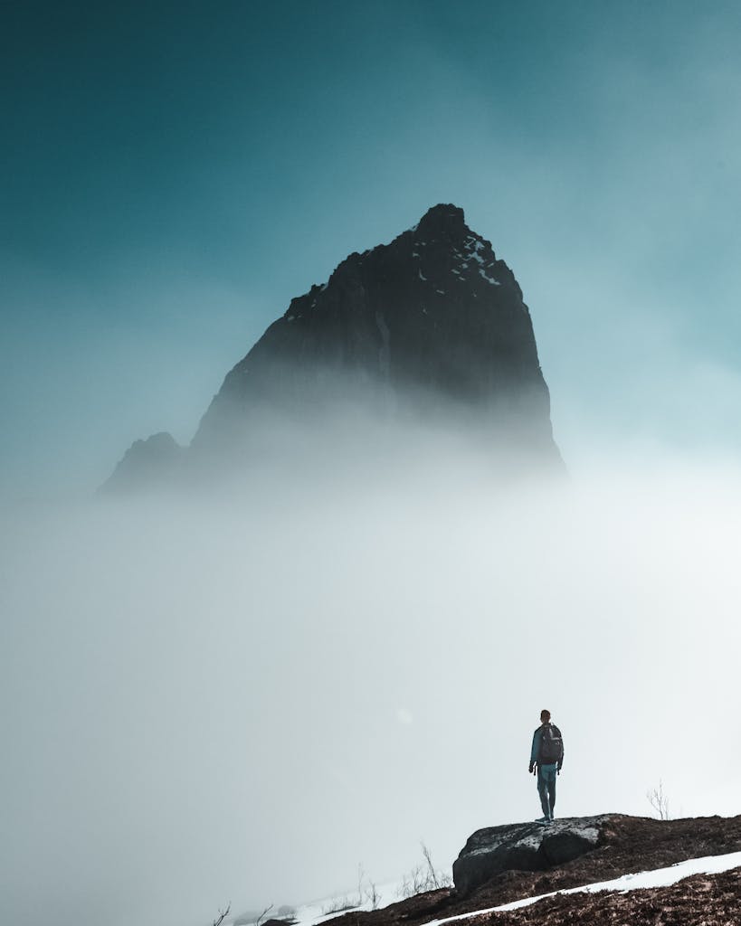 Man stands on rocky terrain surrounded by dense fog with towering mountain in background.